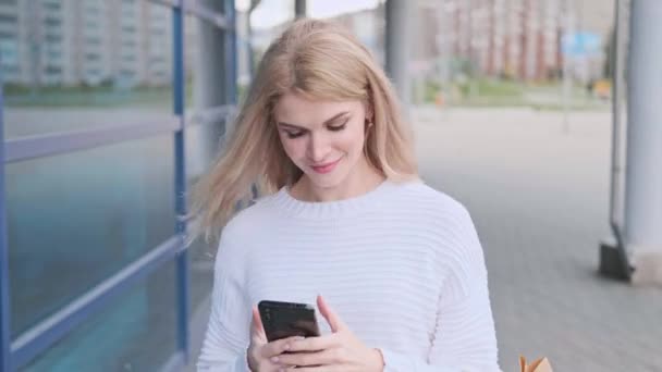 Een jonge mooie blonde vrouw in een witte trui en sneakers loopt door de straat met pakketten van de winkel. Winkelen zwangere vrouw. — Stockvideo