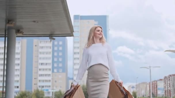 Μια νεαρή όμορφη ξανθιά γυναίκα με ένα λευκό πουλόβερ και αθλητικά παπούτσια περπατά στο δρόμο με πακέτα από το κατάστημα. — Αρχείο Βίντεο