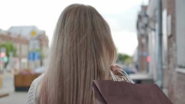 Beyaz ceketli çekici sarışın bir kadın elinde alışveriş torbalarıyla şehrin caddesinde yürüyor. Gülümse ve neşelen..