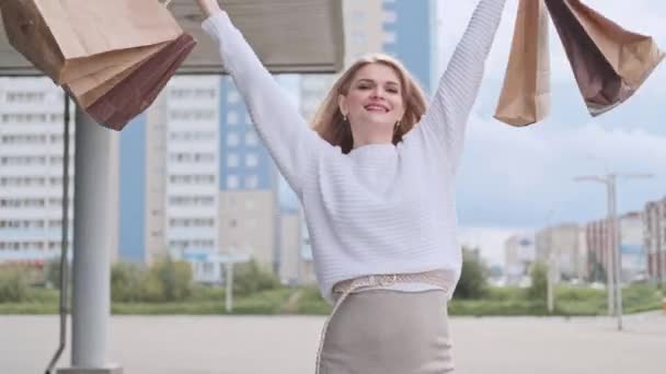 Uma jovem mulher loira bonita em uma camisola branca e tênis caminha pela rua com pacotes da loja . — Vídeo de Stock