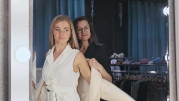 Egy fiatal, vonzó szőke nő egy ruhaboltban felpróbál egy kabátot a tükör előtt.. — Stock videók