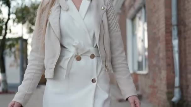 Een jonge aantrekkelijke blonde vrouw in een wit jasje loopt door de straat van de stad met boodschappentassen van de winkel. Glimlachen en vreugde. — Stockvideo