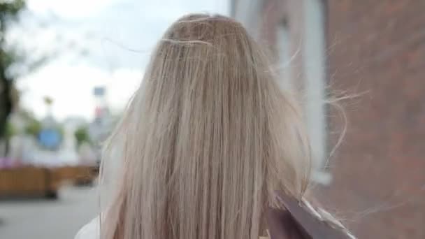 Une jeune jolie femme blonde vêtue d'une veste blanche descend la rue de la ville avec des sacs à provisions du magasin. Souriez et réjouissez-vous . — Video