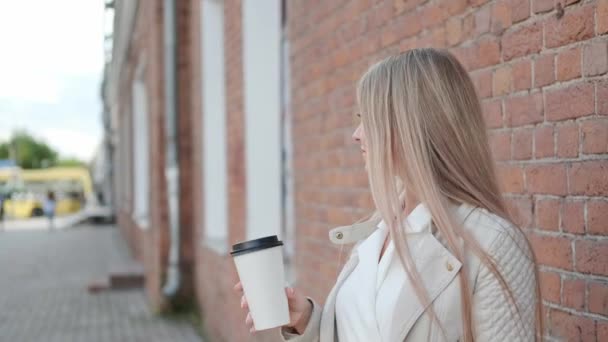 Una joven mujer atractiva rubia al aire libre bebe café de una taza de papel y sostiene paquetes con compras de la tienda . — Vídeos de Stock