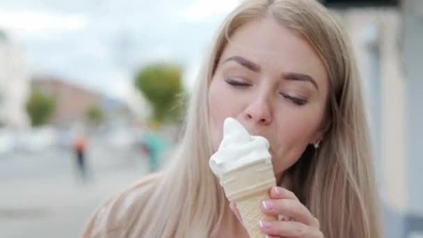 Een jonge aantrekkelijke blonde vrouw eet een witte ijshoorn in een wafelbeker op straat. — Stockvideo