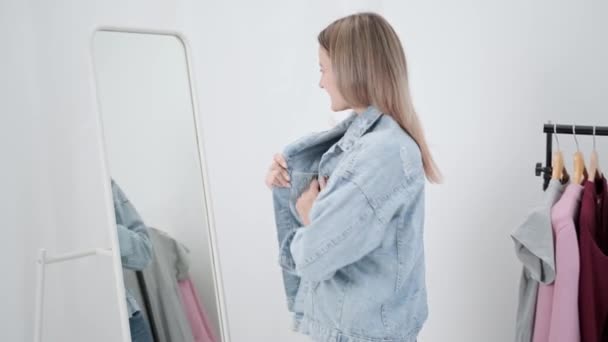 Eine junge attraktive Millennial-Frau in weißem Zimmer probiert an und wählt Kleidung aus. Sieht im Spiegel und vergleicht Outfits. Garderobe des Hauses oder Umkleideraum im Geschäft. — Stockvideo