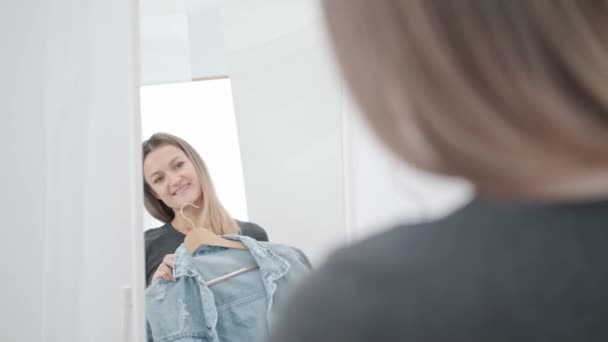 Uma jovem mulher milenar atraente na sala branca experimenta e escolhe roupas. Olha no espelho e compara roupas. Sala de guarda-roupa da casa ou sala de montagem na loja . — Vídeo de Stock