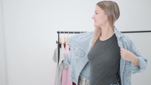 A young attractive millennial woman in white room tries on and chooses clothes. Looks in mirror and compares outfits. Wardrobe room of house or fitting room in store. — Stock Video