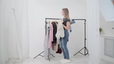 Beyaz odadaki genç ve çekici bir kadın deneyip kıyafet seçiyor. Aynaya bakar ve kıyafetleri karşılaştırır. Soyunma odası ya da soyunma odası..