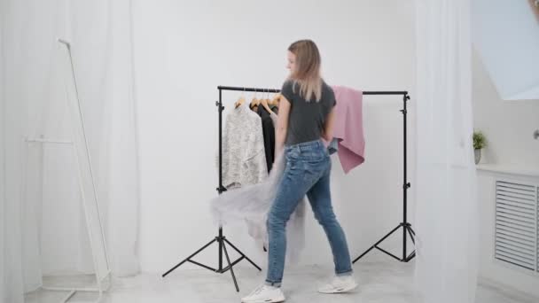 Egy fiatal, vonzó Y-generációs nő fehér szobában felpróbálja, és ruhákat választ. Nézz tükörbe és hasonlítsd össze a ruhákat. Ruhaszekrény szoba ház vagy próbaterem a raktárban. — Stock videók