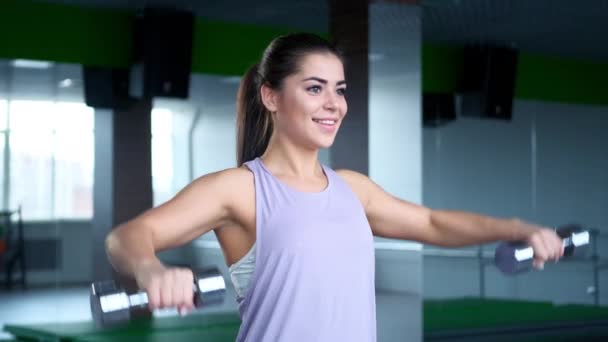 Une belle jeune femme brune sportive dans les trains de vêtements de sport avec des haltères dans la salle de gym . — Video