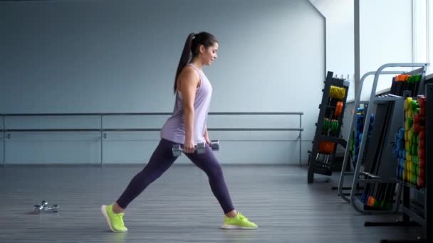 Une belle jeune femme brune sportive dans les trains de vêtements de sport avec des haltères dans la salle de gym . — Video