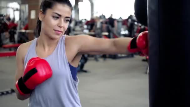 Atletische jonge brunette vrouw in sportkleding en rode bokshandschoenen treinen hobbels op een bokszak in een fitnessruimte. — Stockvideo