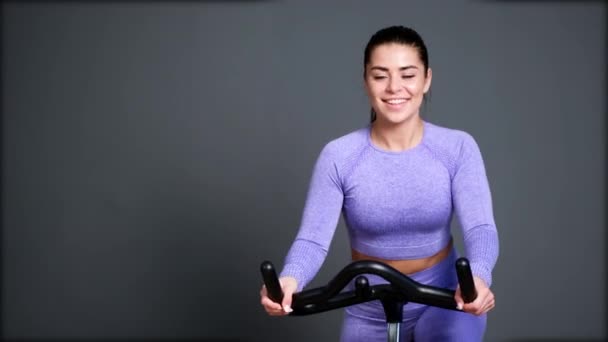 Une belle jeune femme brune sportive en vêtements de sport s'entraîne sur un sycle dans la salle de gym contre le mur gris. — Video