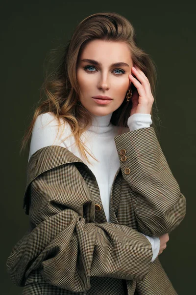 Een jonge mooie blonde vrouw in een witte coltrui en een mans jasje poseert op een groene olijf achtergrond in de studio. — Stockfoto