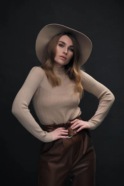 A young sensual sexy woman with a beautiful face and clear natural skin poses in the studio against a black background. Beige slinky sweater and hat with fields. — Stock Photo, Image