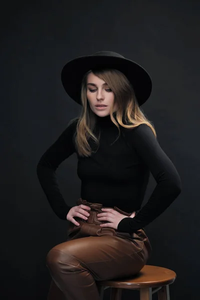 Portrait of young beautiful blond woman with light makeup and blue eyes touching her face. Dark background. Black tight sweater and hat with fields. — Stock Photo, Image
