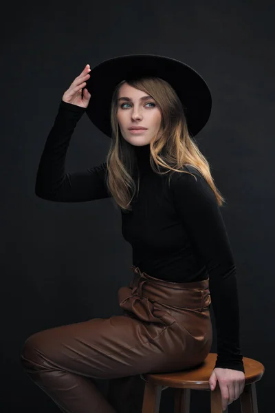 Portrait of young beautiful blond woman with light makeup and blue eyes touching her face. Dark background. Black tight sweater and hat with fields. — Stock Photo, Image