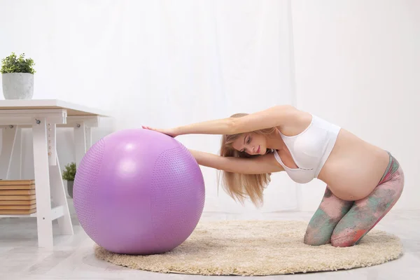 Eine junge schöne schwangere Kaukasierin sitzt auf dem weichen Teppich des Hauses. Er betreibt Fitness und macht Übungen. Sportbekleidung. — Stockfoto