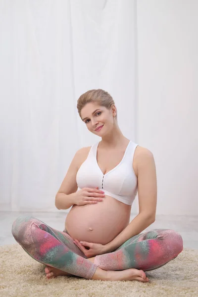 Een jonge mooie zwangere blanke vrouw zit op het zachte tapijt van het huis. Hij is bezig met fitness en oefeningen. Sportkleding. — Stockfoto