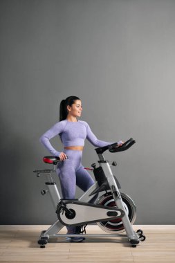 Spor kıyafetleri içinde güzel bir esmer kadın spor salonunda gri duvara karşı bisiklet sürüyor..