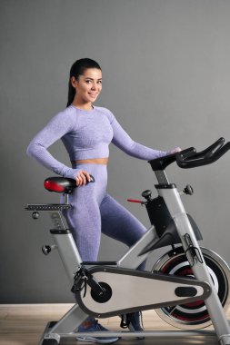 Spor kıyafetleri içinde güzel bir esmer kadın spor salonunda gri duvara karşı bisiklet sürüyor..