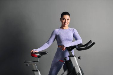 Spor kıyafetleri içinde güzel bir esmer kadın spor salonunda gri duvara karşı bisiklet sürüyor..