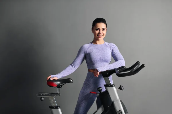 Eine schöne, athletische junge brünette Frau in Sportbekleidung trainiert auf einem Fahrrad in der Turnhalle gegen die graue Wand. — Stockfoto