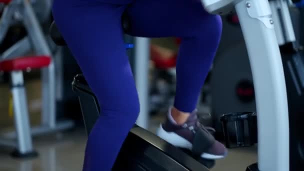 Eine schöne athletische junge brünette Frau in Sportbekleidung im Fitnessstudio ist mit einem Fahrrad verlobt. — Stockvideo