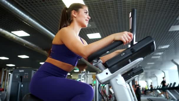 Μια όμορφη αθλητική νεαρή μελαχρινή γυναίκα σε αθλητικά ρούχα στο γυμναστήριο ασχολείται με ένα ποδήλατο. — Αρχείο Βίντεο