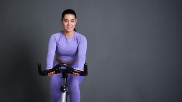 Eine schöne, athletische junge brünette Frau in Sportbekleidung trainiert auf einem Fahrrad in der Turnhalle gegen die graue Wand. — Stockvideo