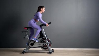 Spor kıyafetleri içinde güzel bir esmer kadın spor salonunda gri duvara karşı bisiklet sürüyor..