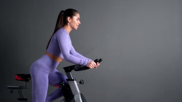 Una bella giovane donna bruna atletica in abbigliamento sportivo si allena su un sycle in palestra contro il muro grigio. — Video Stock