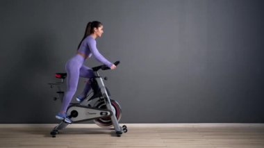 Spor kıyafetleri içinde güzel bir esmer kadın spor salonunda gri duvara karşı bisiklet sürüyor..