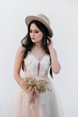 Gelin portresi. Uzun siyah saçlı, örgülü, çekici, esmer bir kadın. Beyaz gelinlik ve bej şapka. Kırsal stil, buğday buketi..