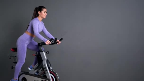 Una hermosa joven morena atlética en ropa deportiva entrena en un sycle en el gimnasio contra la pared gris. — Vídeo de stock