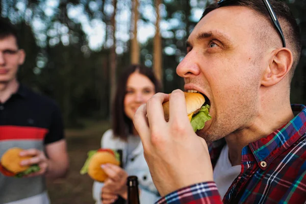 Un grupo de amigos descansa y se relaja en la naturaleza. La gente come hamburguesas, bebe cerveza, se comunica y ríe . — Foto de Stock