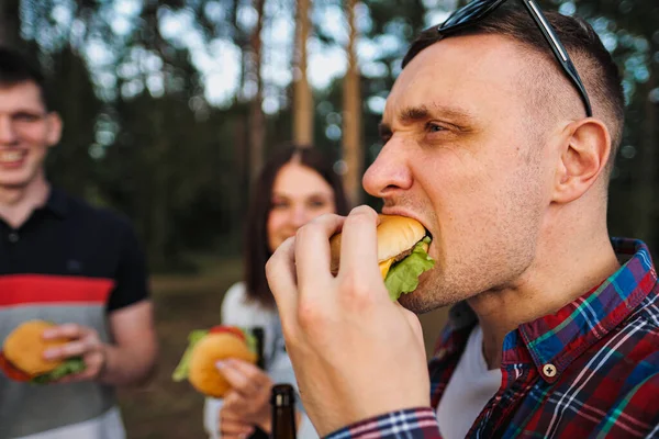 Un grupo de amigos descansa y se relaja en la naturaleza. La gente come hamburguesas, bebe cerveza, se comunica y ríe . — Foto de Stock