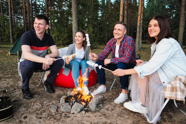 A group of friends relax in a forest camp. Men and women prepare a marshmallow on a bonfire. A party in nature.