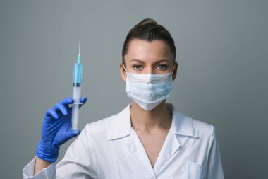Beyaz cüppeli ve maskeli bir kadın doktor elinde bir şırınga tutuyor..