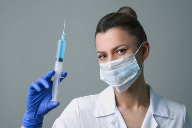 Beyaz cüppeli ve maskeli bir kadın doktor elinde bir şırınga tutuyor..