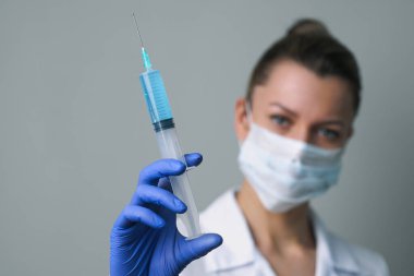 Beyaz cüppeli ve maskeli bir kadın doktor elinde bir şırınga tutuyor..