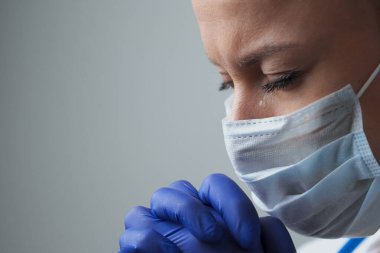 Beyaz cüppeli, maskeli ve eldivenli bir kadın doktor. Yüzü yakın plan. Doktor ağlar ve dua eder. Gözyaşları. Salgın ve virüs salgını. Coronavirus covid-19.