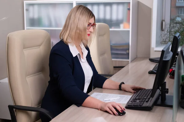 Mladá blondýna s brýlemi je kancelářská pracovnice. Žena sedí u stolu a pracuje u počítače. — Stock fotografie