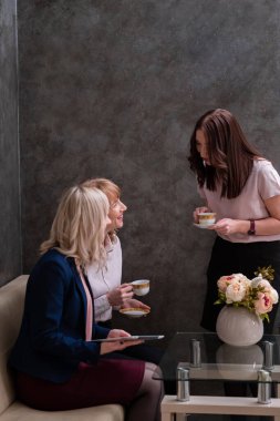 Üç genç kadın iş yerinde sosyalleşiyor ve çay içiyor. Gri duvar.