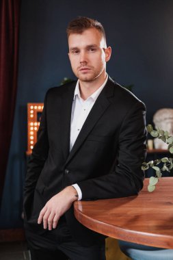 Yakışıklı, genç, sakallı, takım elbiseli ve gömlekli sarışın bir adam. Masaya oturur, dergi okur..