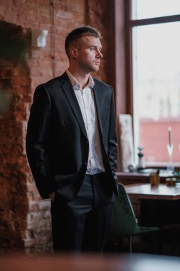 Yakışıklı, genç, sakallı, takım elbiseli ve gömlekli sarışın bir adam. Masaya oturur, dergi okur..