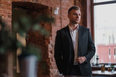 Yakışıklı, genç, sakallı, takım elbiseli ve gömlekli sarışın bir adam. Masaya oturur, dergi okur..