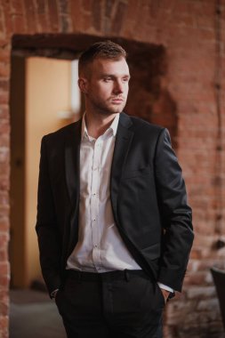 Yakışıklı, genç, sakallı, takım elbiseli ve gömlekli sarışın bir adam. Masaya oturur, dergi okur..