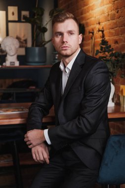 Yakışıklı, genç, sakallı, takım elbiseli ve gömlekli sarışın bir adam. Masaya oturur, dergi okur..